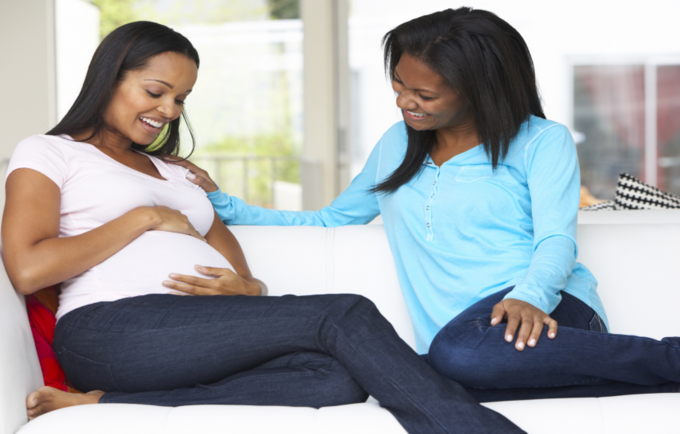 Image showing two (2) ladies sitting on a couch with one of the ladies being pregnant and rubbing tummy