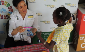 Picture showing a staff member of Vision 2030 Jamaica handing a pink pen to a little girl at the UNFPA World Population Day 2017 Commemoration Ceremony that was held in Santa Cruz, St. Elizabeth on July 11, 2017