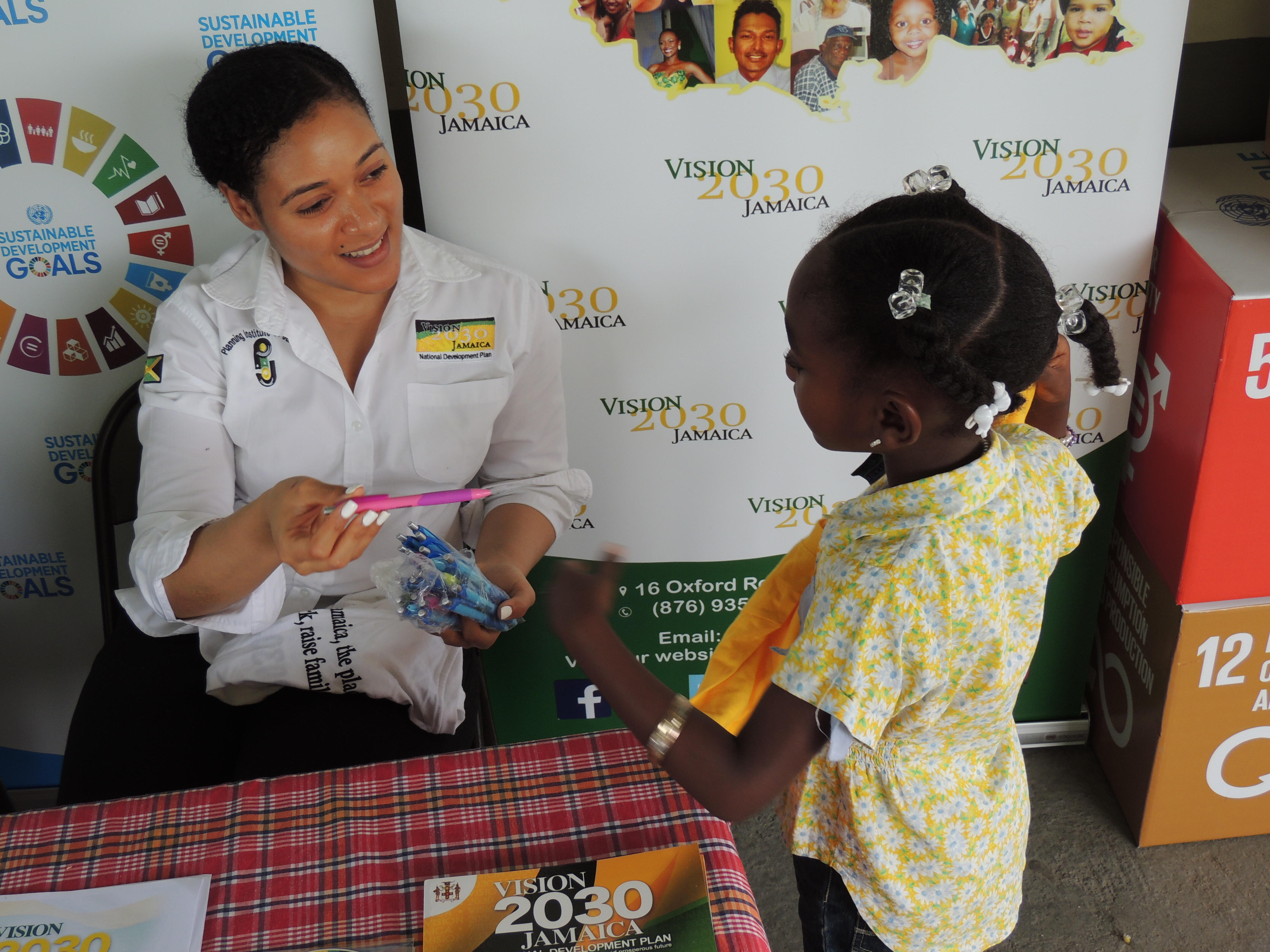 Picture showing a staff member of Vision 2030 Jamaica handing a pink pen to a little girl at the UNFPA World Population Day 2017 Commemoration Ceremony that was held in Santa Cruz, St. Elizabeth on July 11, 2017