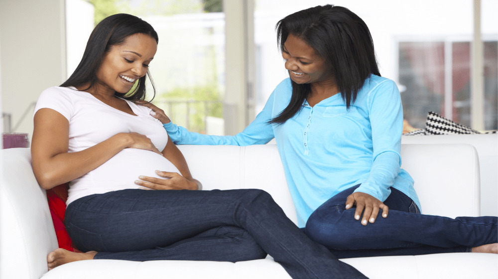 Image showing two (2) ladies sitting on a couch with one of the ladies being pregnant and rubbing tummy